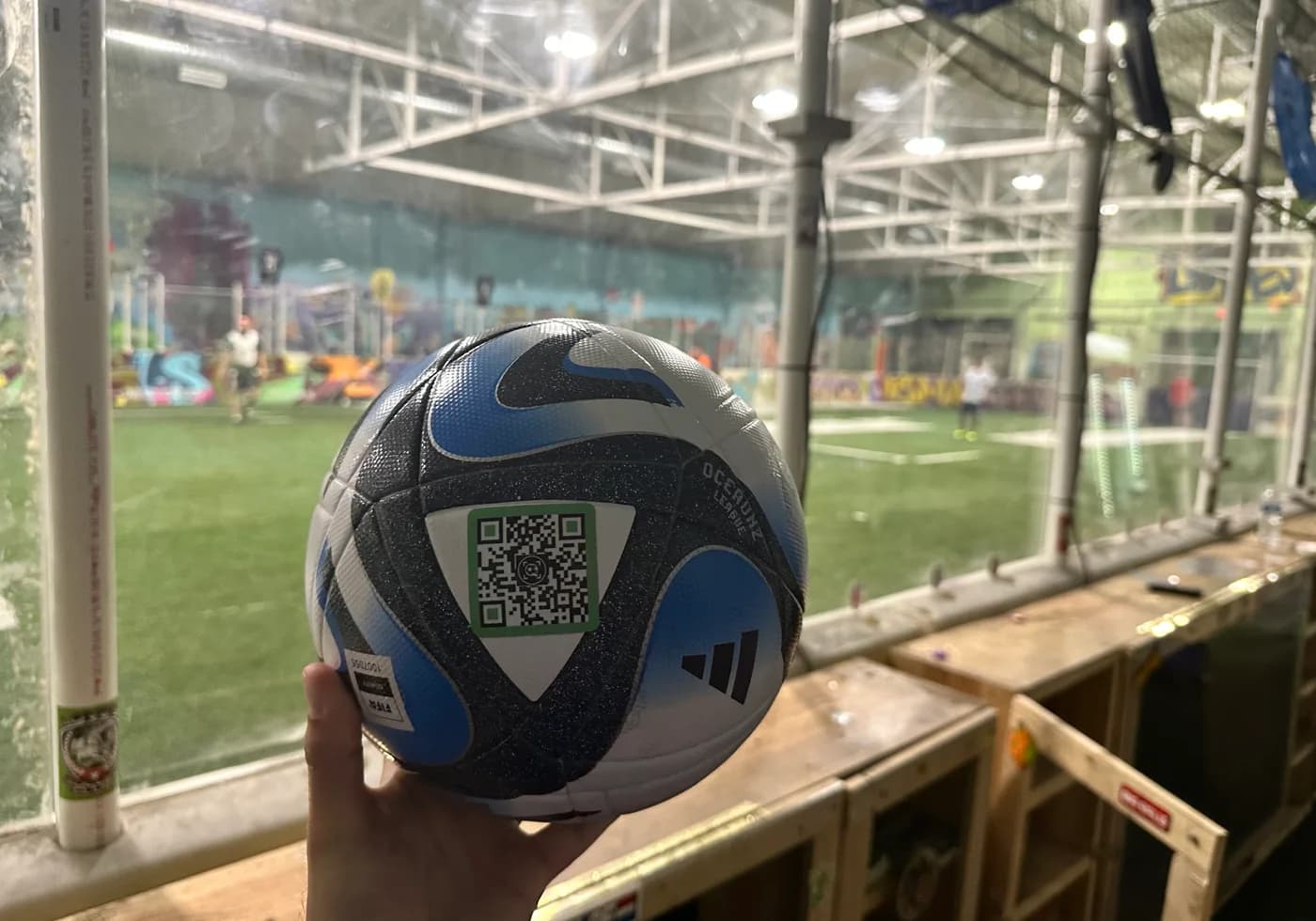 A blue SoccQR-tagged soccer ball held beside an indoor practice field